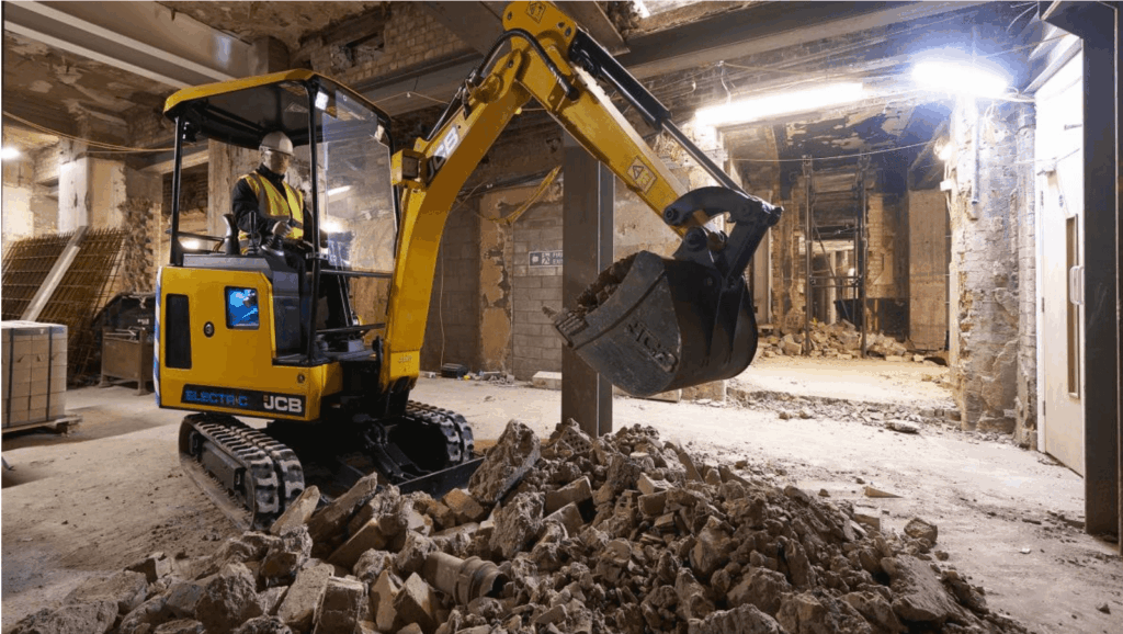 Electric mini excavator carrying out indoor demolition work in a basement environment, breaking up concrete and clearing rubble.