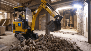 Electric mini excavator carrying out indoor demolition work in a basement environment, breaking up concrete and clearing rubble.