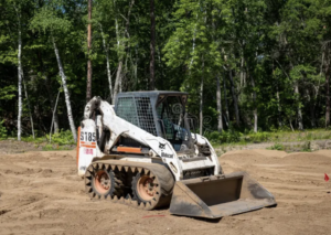 Bobcat S185 skid steer loader with a front bucket parked on a freshly graded dirt construction site surrounded by green trees.